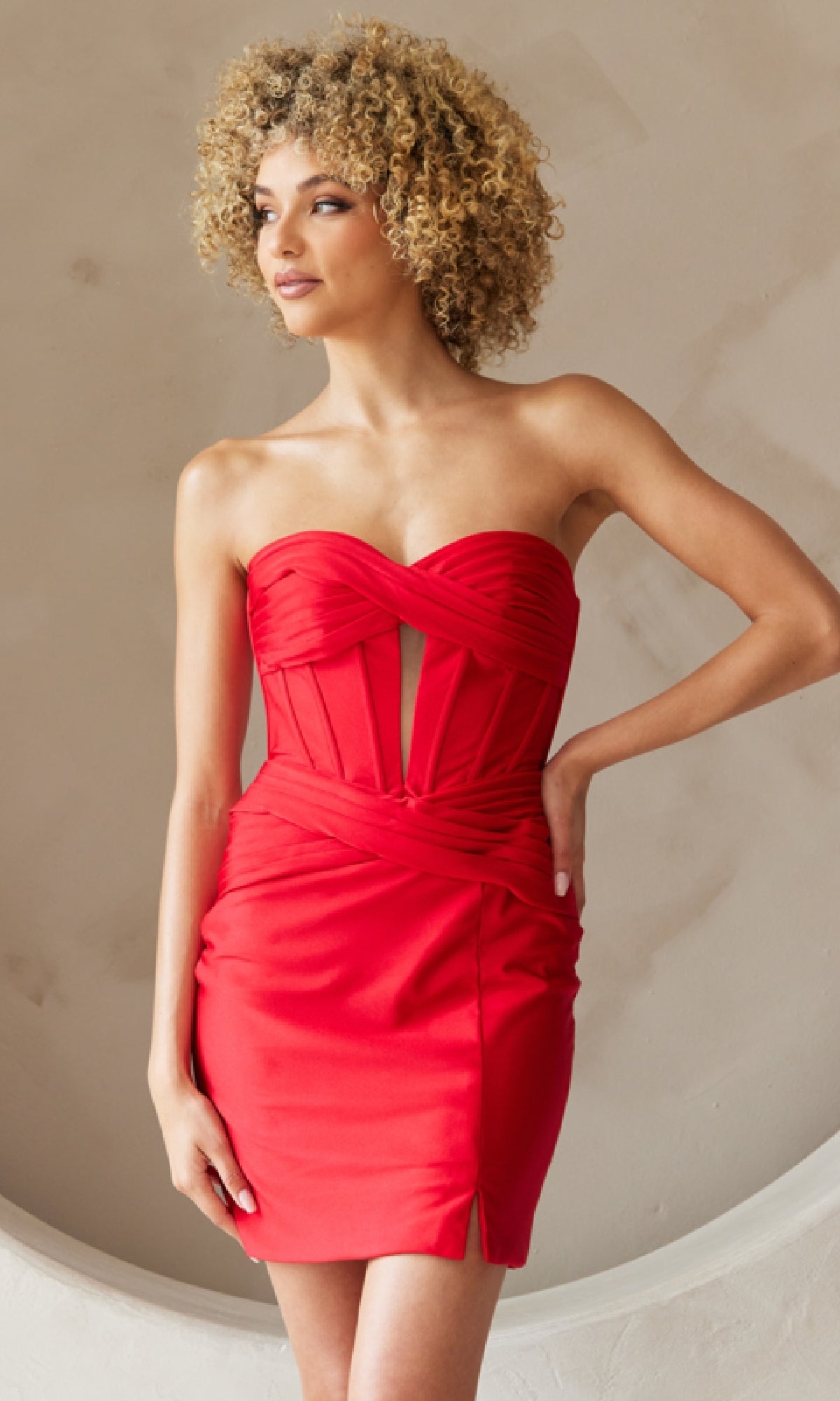 A woman with curly hair wears the Cut-Out Strapless Short Hoco Dress AG0114S, a fitted red cocktail dress with a corset-style bodice, posing against a neutral textured background.